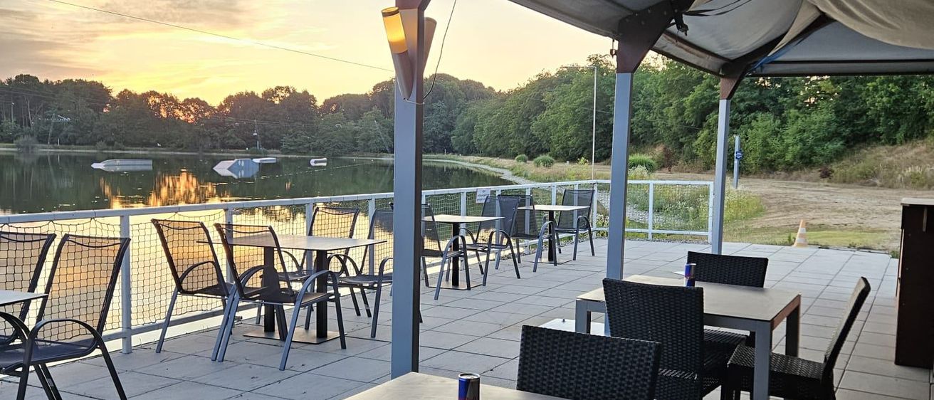 Eine Restaurantterrasse am See mit Tischen und Stühlen unter einem Baldachin, die Sonne geht in der Ferne unter.