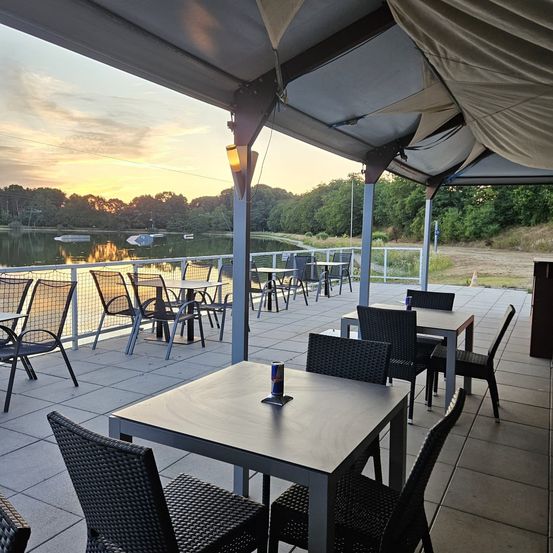 Ein Terrassenbereich mit Tischen und Stühlen in der Nähe eines Sees, der von einem Vordach bedeckt ist. Ein Sonnenuntergang ist im Hintergrund sichtbar, mit Bäumen und einem Boot auf dem Wasser.