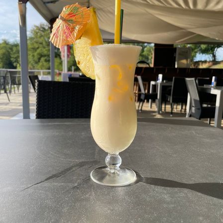 Ein hohes Glas mit einem weißen Getränk, einem gelben Strohhalm und einer Ananasscheibe. Es steht auf einem Tisch in einem Restaurant mit Stühlen und Tischen.