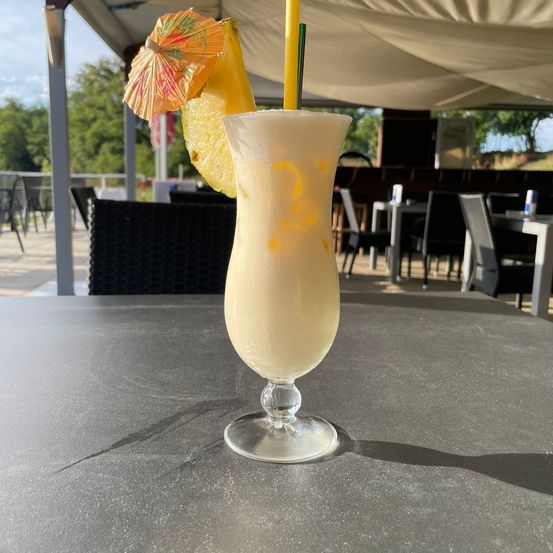 Ein hohes Glas mit einem weißen Getränk, einem gelben Strohhalm und einer Ananasscheibe. Es steht auf einem Tisch in einem Restaurant mit Stühlen und Tischen.