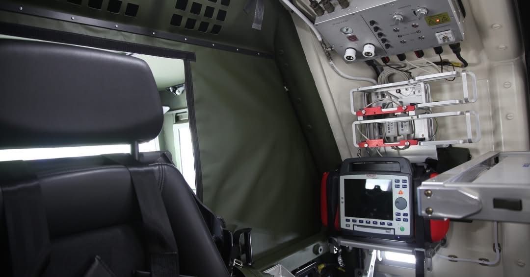 Das Innere eines Militärfahrzeugs zeigt einen schwarzen Ledersitz, ein elektronisches medizinisches Gerät und verschiedene Bedienfeldern mit Tasten und Anzeigen.