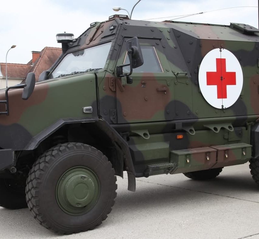 Ein gepanzertes Fahrzeug mit getarntem Äußeren steht vor einem Gebäude. Das Fahrzeug hat ein rotes Kreuzsymbol auf der Vorderseite und ein großes Fenster.