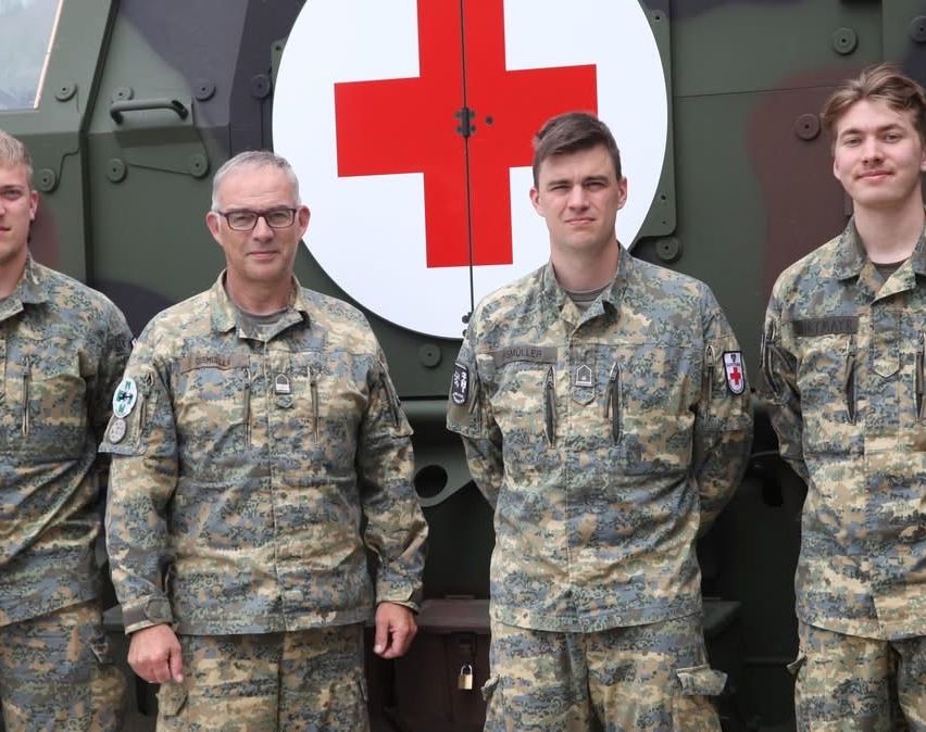 Vier Soldaten stehen vor einem Militärfahrzeug mit einem roten Kreuz-Symbol darauf. Sie tragen Camouflage-Uniformen, und der Mann in der Mitte trägt eine Brille.