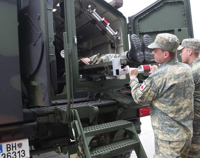 Zwei Soldaten in Tarnuniformen helfen einem Soldaten, der auf einer Trage in einem Militärfahrzeug liegt. Ein Soldat hält eine rot-weiße Flasche, möglicherweise Sauerstoff, während der andere ein rotes Kreuz am Arm hat.