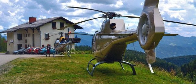 Zwei Hubschrauber sind auf einem grasbewachsenen Hügel geparkt, mit einem Haus und geparkten Autos. Eine Person steht in der Nähe des Hubschraubers.