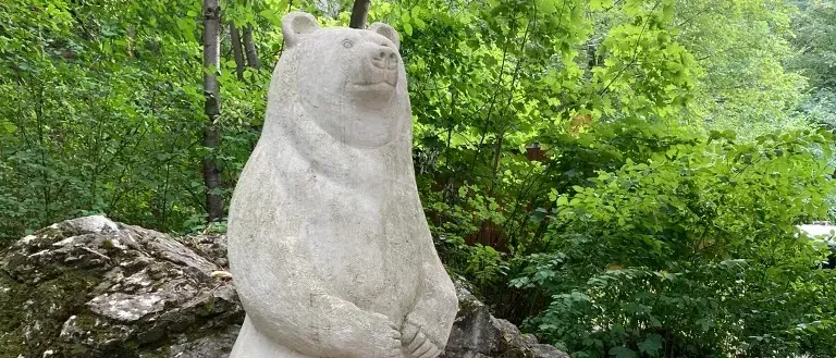 Eine Steinbärenstatue steht auf einem Felsen in einem üppigen Wald mit einem Informationsschild in der Nähe.