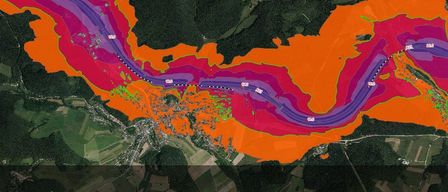 Ein Satellitenbild zeigt einen mäandernden Fluss mit Überschwemmungsgebieten in Lila- und Orangetönen. Grüne Markierungen zeigen städtische Gebiete entlang der Flussufer an.