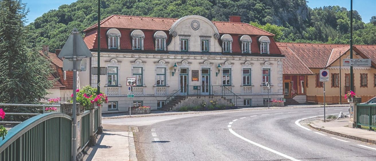 Ein malerisches Gebäude mit rotem Dach und vielen Fenstern steht an einer Straßenecke, mit einem Berg und üppigem Grün im Hintergrund.