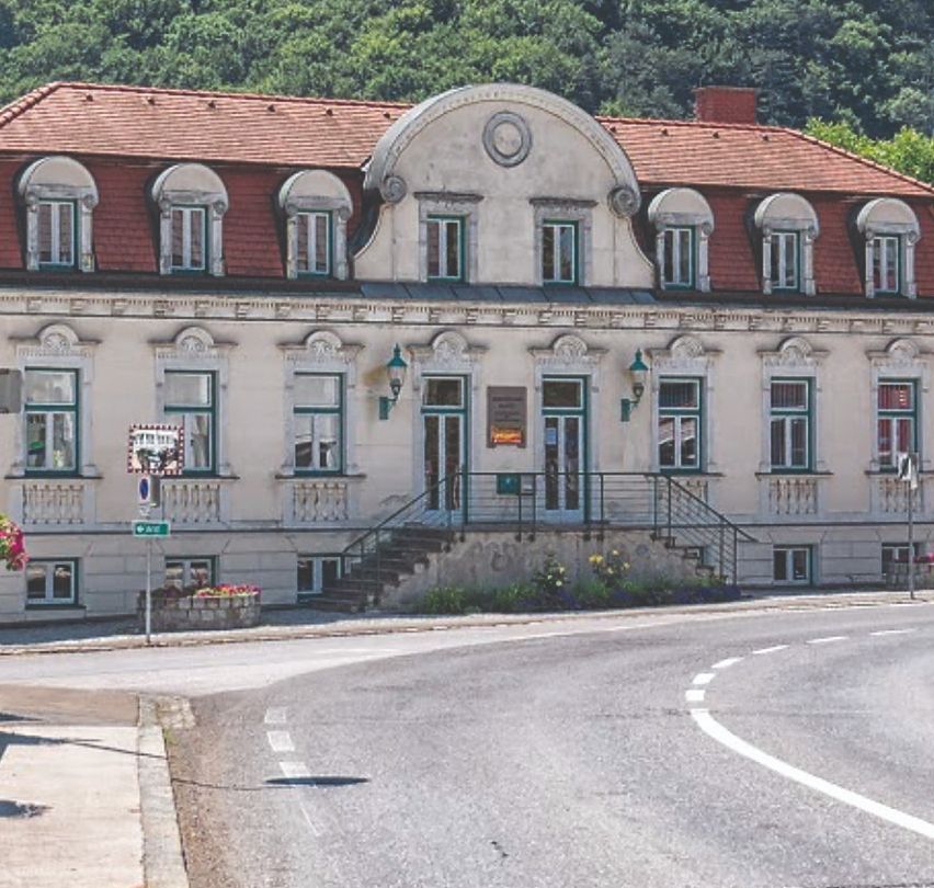 Ein malerisches Gebäude mit rotem Dach und vielen Fenstern steht an einer Straßenecke, mit einem Berg und üppigem Grün im Hintergrund.