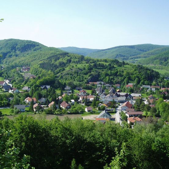 Ein malerisches Dorf eingebettet zwischen üppigen grünen Hügeln, mit Häusern unterschiedlicher Größe und Farbe, umgeben von dichten Bäumen und einem klaren blauen Himmel.