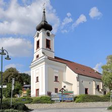 Eine weiße Kirche mit schwarzem Turm steht unter blauem Himmel. Sie hat eine Uhr und Kreuze oben. In der Nähe gibt es Straßenschilder und eine Straßenlampe.