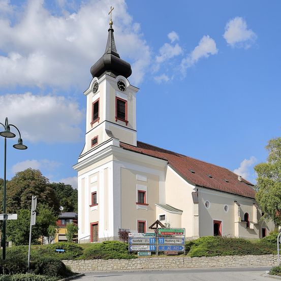 Eine weiße Kirche mit schwarzem Turm steht unter blauem Himmel. Sie hat eine Uhr und Kreuze oben. In der Nähe gibt es Straßenschilder und eine Straßenlampe.