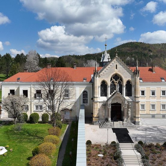 Ein Blick von oben auf eine große, prächtige Kirche mit rotem Dach und einer Statue über dem Eingang. Das Gebäude ist von einem gepflegten Garten umgeben, der von einem Kiesweg zum Eingang führt.