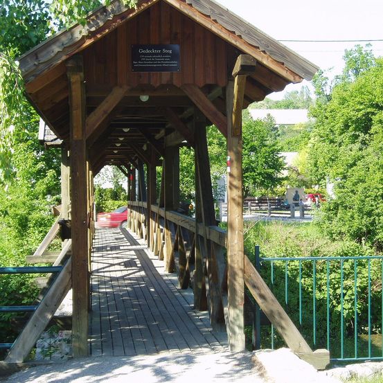 Eine hölzerne überdachte Brücke mit einem Schild, das 'Gedeckter Steg' besagt, ist zu sehen. Die Brücke befindet sich über einem Fluss, und ein roter Wagen ist in der Nähe geparkt.