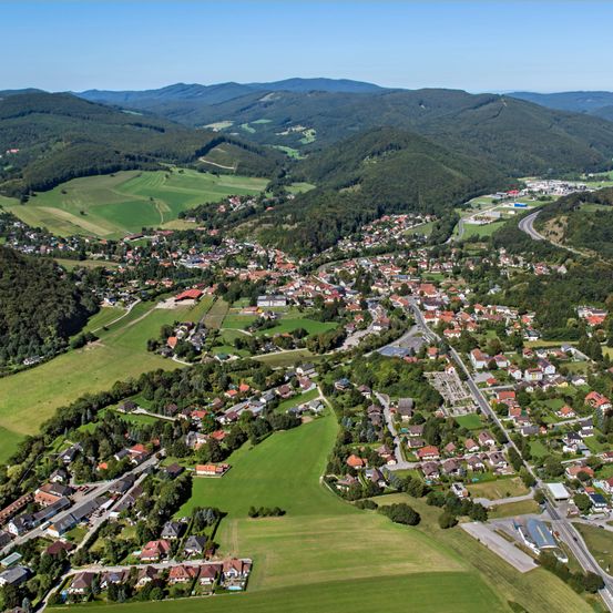 Eine Luftaufnahme einer Stadt, umgeben von grünen Feldern und Hügeln. Die Stadt hat viele Häuser, einige mit roten Dächern, und liegt eingebettet zwischen üppigen Wäldern. Eine Straße schlängelt sich durch die Stadt und verbindet sie mit den umliegenden Gebieten.