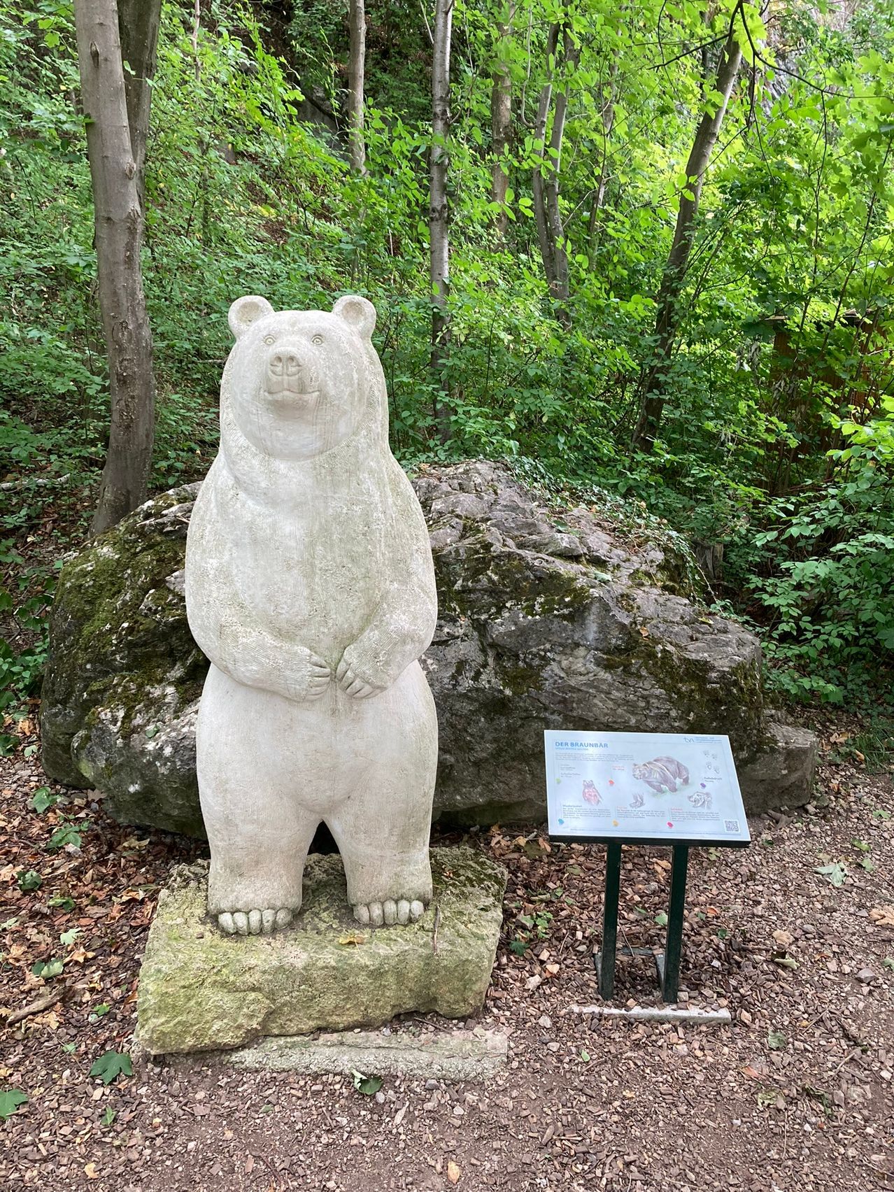 Eine Steinstatue eines Bären steht auf einem Felsen in einem üppigen grünen Wald, mit einem Schild in der Nähe, das über den Bären informiert.