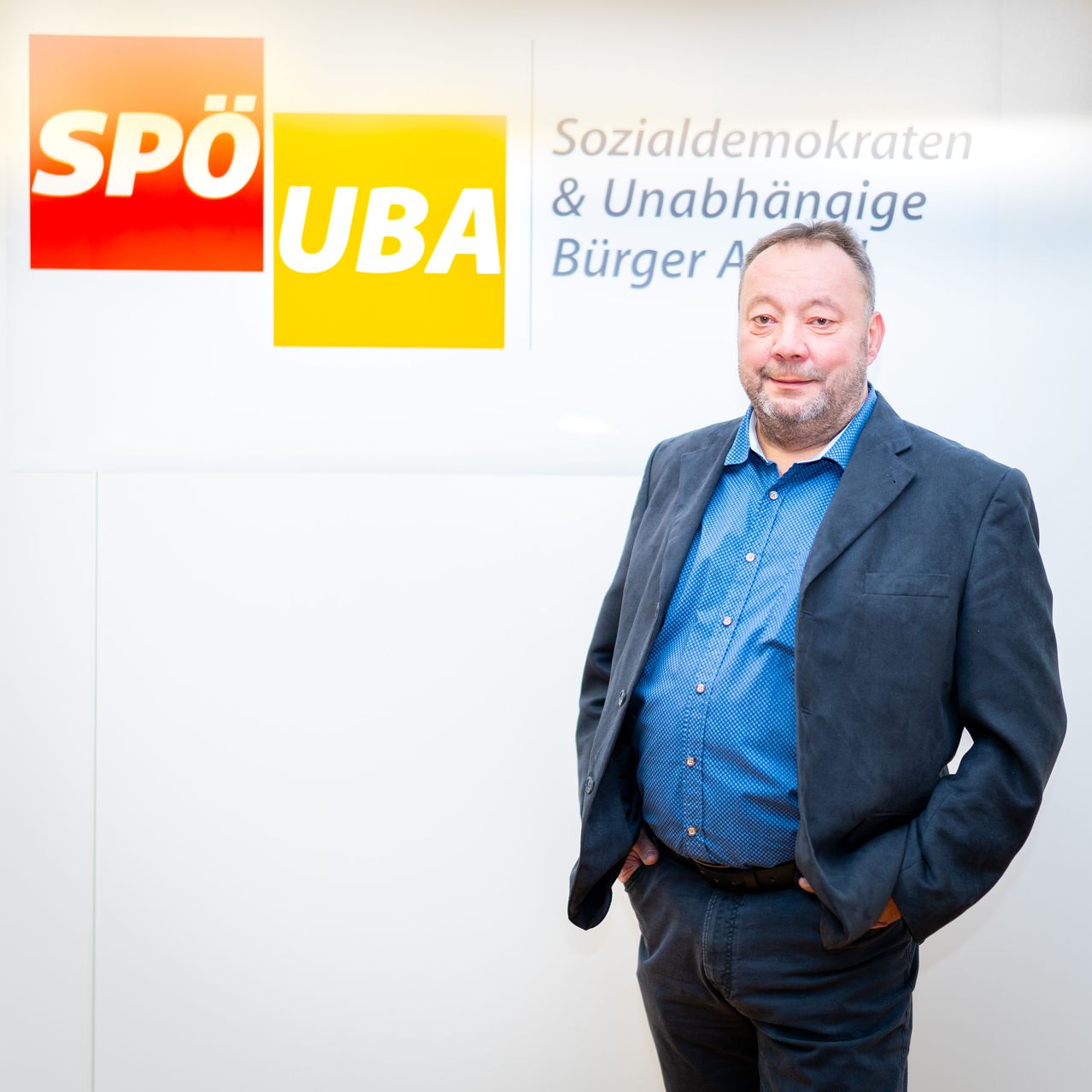 Ein Mann steht vor einer Wand mit politischen Logos. Er trägt ein blaues Hemd und einen grauen Anzug. Seine Hände sind in den Taschen.