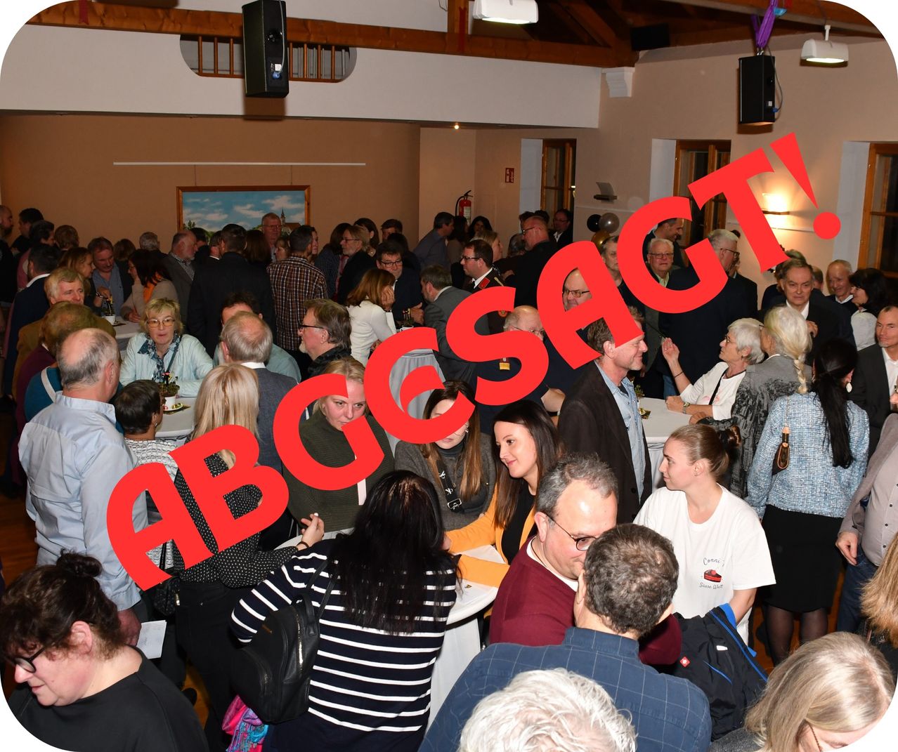 Eine große Menschenmenge in einem Saal mit Tischen und Stühlen. Das Wort 'ABCGCSAGT!' ist in Rot auf dem Bild geschrieben.