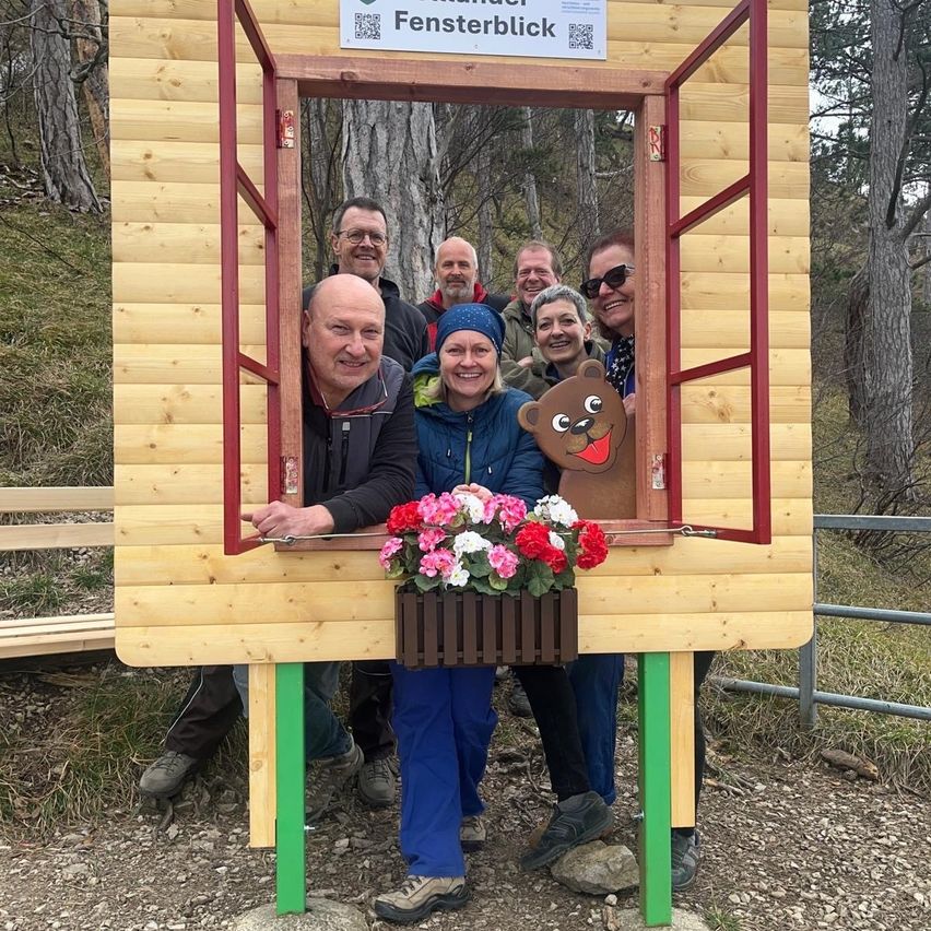 A group of people pose next to a wooden booth with flowers and a sign that says Fensterblick.