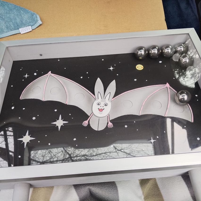A table with a glass tray featuring a cartoon bat on a night sky background with stars. Silver balls are scattered on the tray.