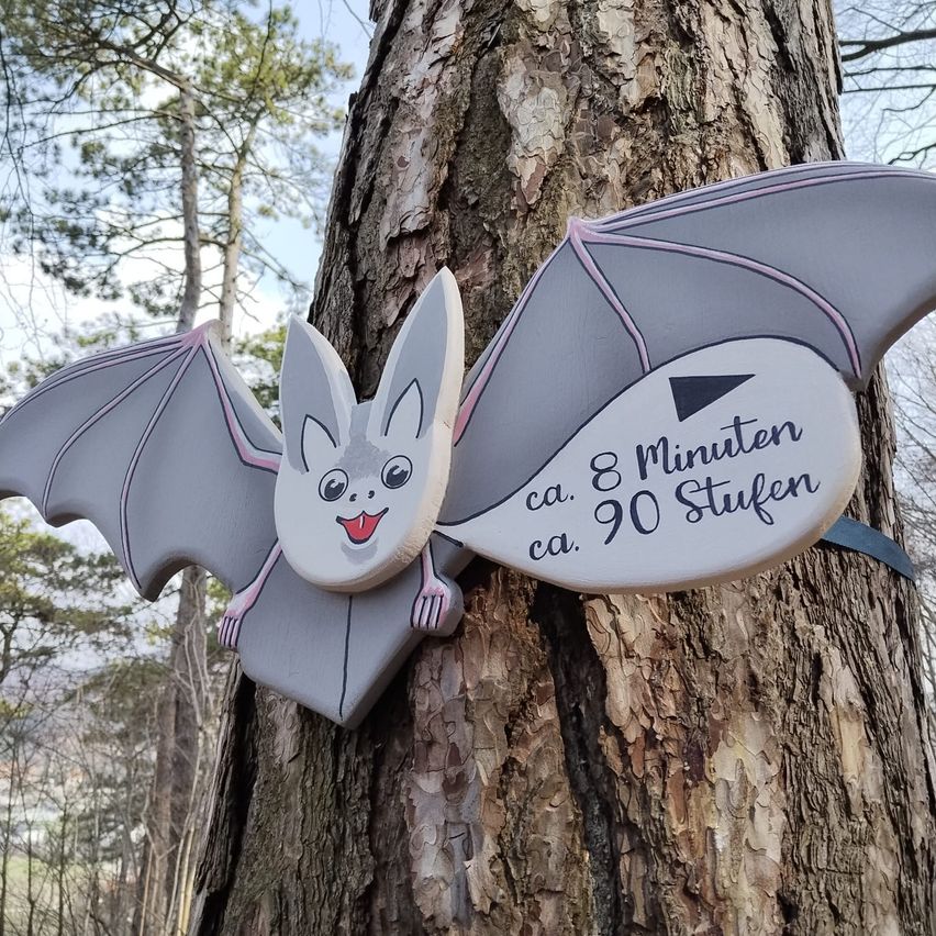 A bat-shaped sign is affixed to a tree trunk, showing directions in gray and white. It reads approximately 8 minutes and 90 steps.