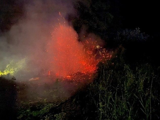 Ein Feuer brennt in der Nacht und beleuchtet die dunkle Umgebung mit roten und orangefarbenen Flammen. Rauch und Funken steigen in die Luft auf.