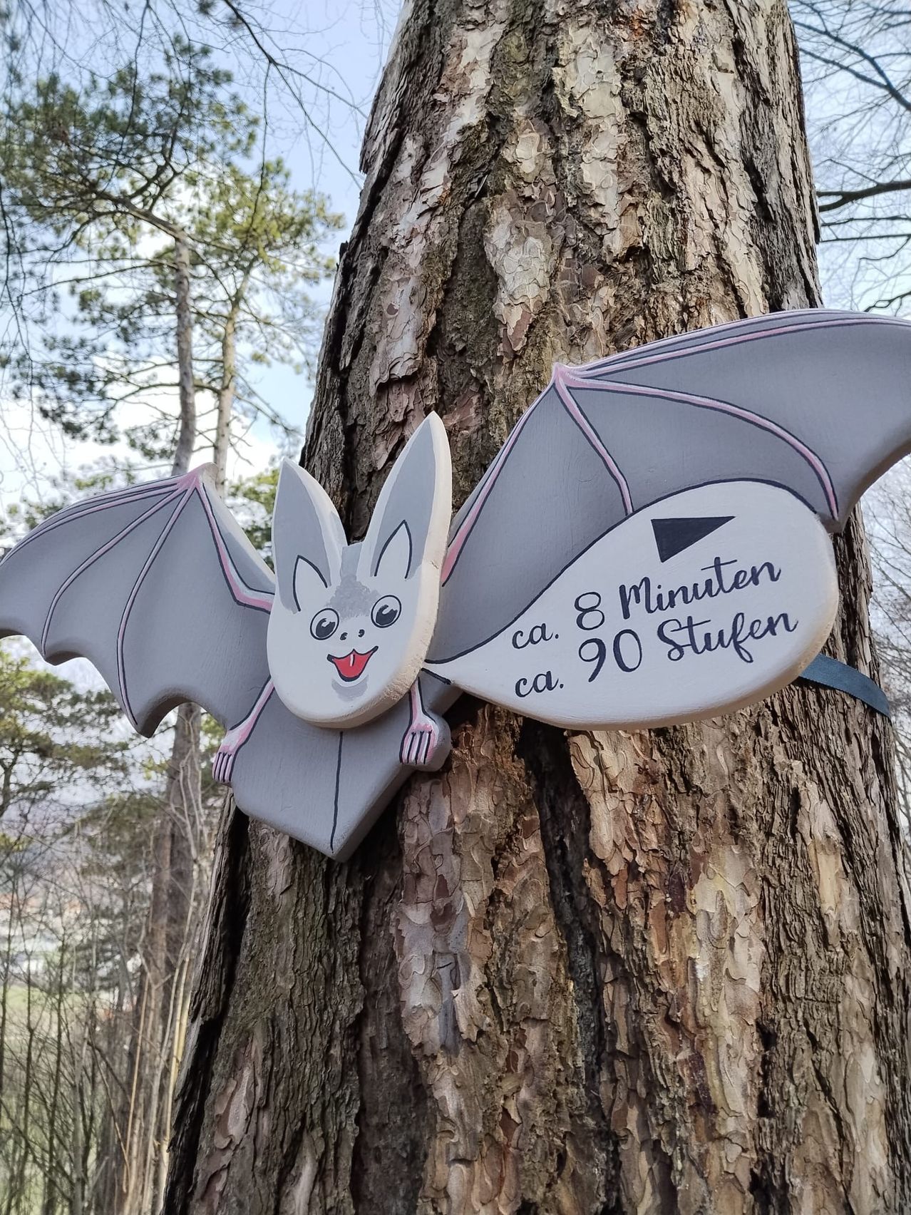 Ein hölzerne Fledermaus-Schild hängt an einem Baum. Es hat ein lächelndes Gesicht, graue Flügel und Text, der 'ca. 8 Minuten, ca. 90 Stufen' lautet. Der Baum ist von vielen Bäumen umgeben, und das Gebiet ist mit Gras bedeckt.