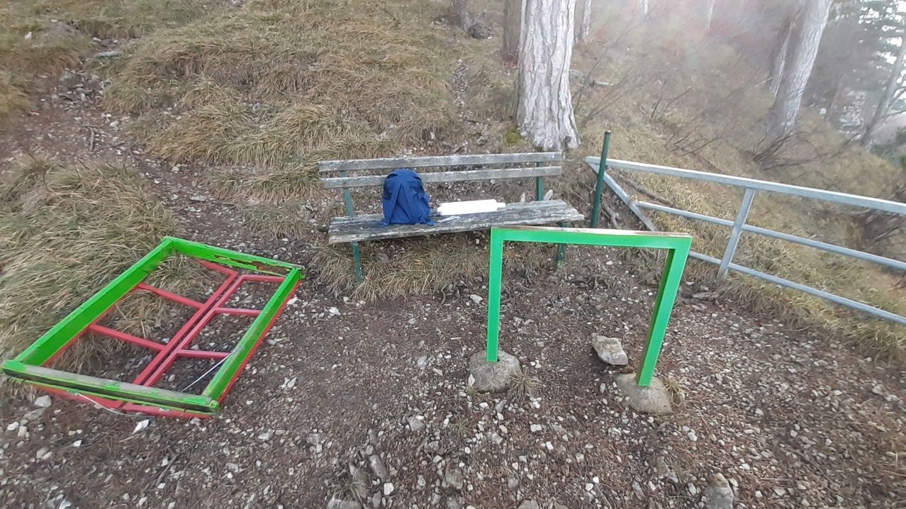 Eine Holzbank steht auf einem felsigen Hang mit einem blauen Rucksack und einem weißen Gegenstand darauf. Grüne Metallpfosten stehen auf dem Boden, und ein rot-grünes Metallobjekt liegt links.