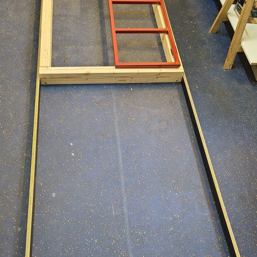 Ein hölzernes Gebilde, das einem Türrahmen ähnelt, mit rotem Rand, steht auf einem blauen Teppichboden. Der Holzrahmen hat zwei quadratische Fenster. Ein hölzernes Regal ist auf der rechten Seite sichtbar.