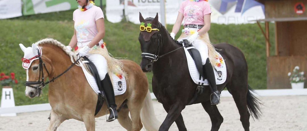 Zwei Mädchen reiten Pferde auf einer Sandbahn mit Gras dahinter. Beide tragen Helme, Sonnenbrillen und Stiefel. Die Pferde sind mit bunten Bändern geschmückt.