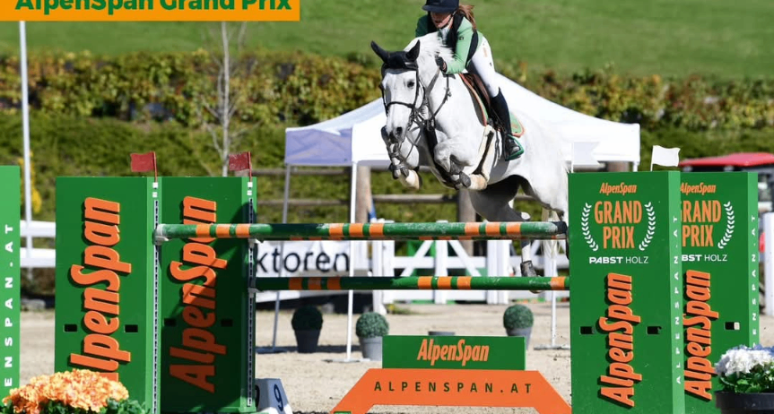 Ein Reiter auf einem weißen Pferd springt ein Hindernis bei einem Wettbewerb mit 'AlpenSpan'-Markenwerbung, auf einem grünen Feld mit einem Zelt im Hintergrund.