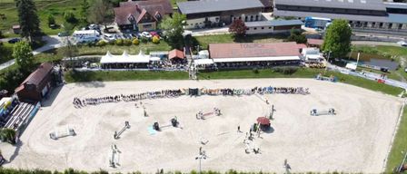 Eine Luftaufnahme eines Pferderennplatzes mit Zuschauern, Zelten und verschiedenen Gebäuden, eingebettet in eine ländliche Landschaft mit grünen Feldern und Bäumen.