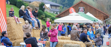 Eine Gruppe von Menschen sitzt auf Heuballen in einem Außenbereich, einige stehen und andere sitzen auf Stühlen. Ein Hund ist anwesend, und Zelte und ein Gebäude befinden sich im Hintergrund.