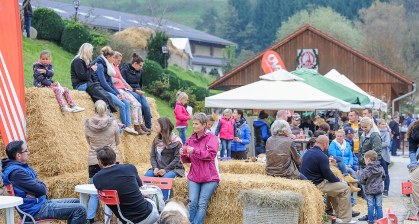 Eine Gruppe von Menschen sitzt auf Heuballen in einem Außenbereich, einige stehen und andere sitzen auf Stühlen. Ein Hund ist anwesend, und Zelte und ein Gebäude befinden sich im Hintergrund.