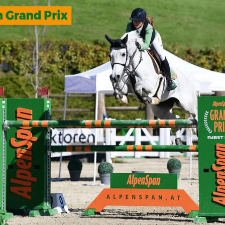Ein Reiter auf einem weißen Pferd springt über eine Hürde mit dem Text 'AlpenSpan' während des 'AlpenSpan Grand Prix.' Ein Zelt ist im Hintergrund.