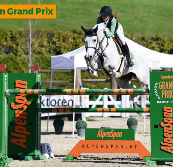 Ein Reiter auf einem weißen Pferd springt über eine Hürde mit dem Text 'AlpenSpan' während des 'AlpenSpan Grand Prix.' Ein Zelt ist im Hintergrund.