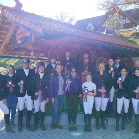 Eine Gruppe von Menschen in Reiterkleidung mit Medaillen posiert für ein Foto vor einer Holzstruktur.