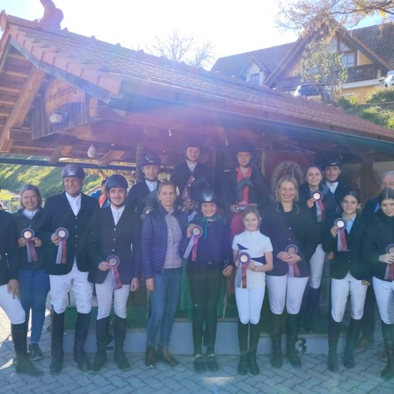 Eine Gruppe von Menschen in Reiterkleidung mit Medaillen posiert für ein Foto vor einer Holzstruktur.
