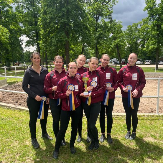 Eine Gruppe von Frauen in roten Jacken steht auf Gras, hält Bänder und Trophäen. Sie lächeln und posieren für ein Foto, mit einem weißen Zaun und Bäumen im Hintergrund.
