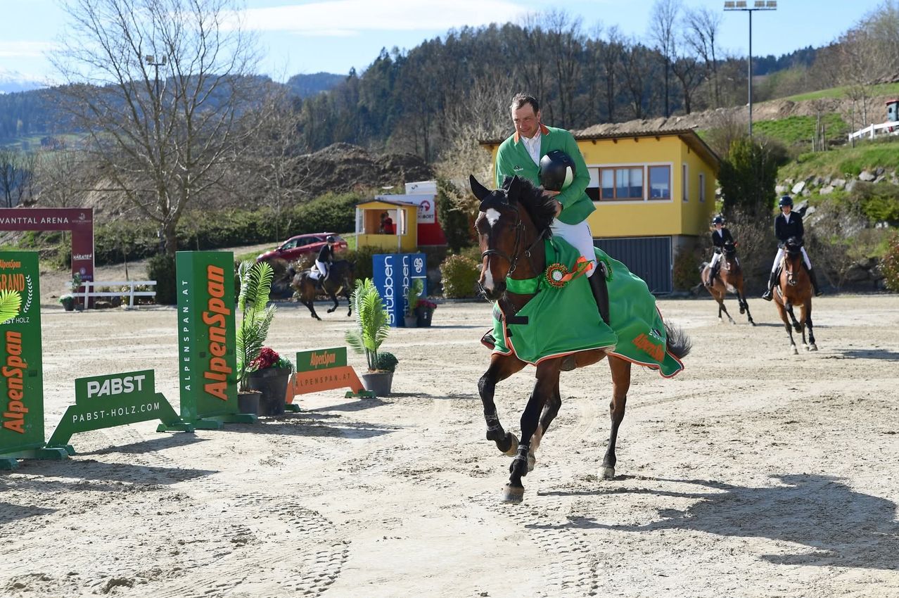 A jockey in a green outfit rides a brown horse in a dirt field with hills in the background.