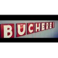 Ein Schild mit dem Wort 'BUCHEREI' an einer weißen Wand. Die Buchstaben sind rot.
