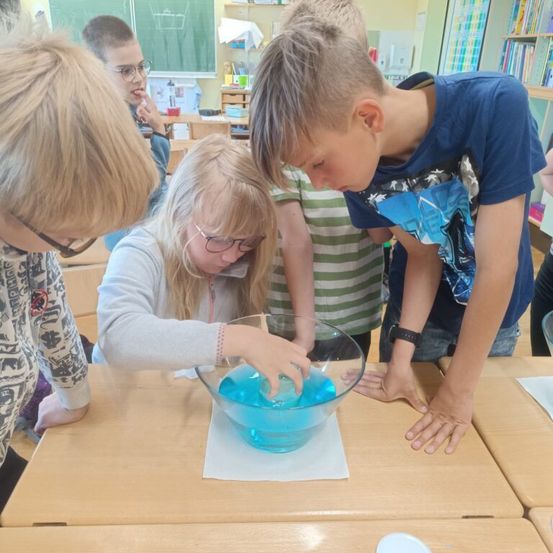 Ein Klassenzimmer mit Schülern, ein Mädchen gießt blaue Flüssigkeit in eine Schüssel auf dem Tisch.