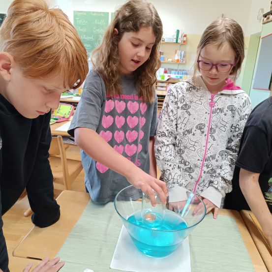Drei Kinder, ein Junge und zwei Mädchen, sind in einem Klassenzimmer. Sie konzentrieren sich auf eine blaue Flüssigkeit in einer Schüssel und führen wahrscheinlich ein Experiment durch.