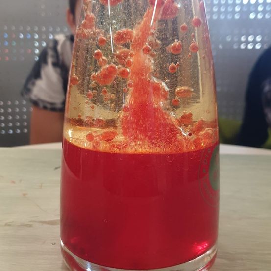 Eine klare Glasflasche mit rotem Inhalt steht auf einem Holztisch. Die Flüssigkeit wird von oben eingegossen und bildet Blasen. Die Flasche ist mit grünem Text beschriftet. Ein Mensch sitzt hinter dem Tisch.