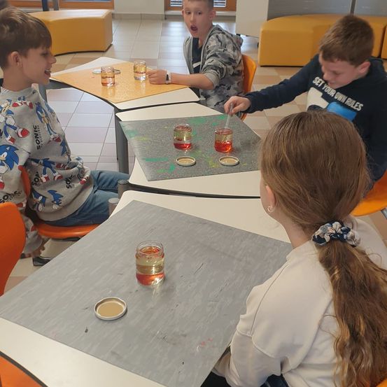 Vier Kinder sitzen um einen Tisch herum, möglicherweise in einem Klassenzimmer. Drei von ihnen schauen auf den Tisch, auf dem sich Gläser mit Flüssigkeit befinden. Ein Kind hält einen Löffel und scheint etwas zu vermischen.