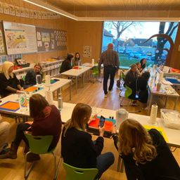 Mehrere Frauen und ein Mann sitzen um Tische in einem Klassenzimmer. Sie beschäftigen sich mit Aktivitäten, und ein Mann geht zwischen den Tischen hindurch. Das Klassenzimmer hat Holzwände und ein großes Fenster mit Blick auf Bäume und geparkte Autos.