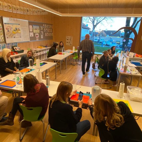Mehrere Frauen und ein Mann sitzen um Tische in einem Klassenzimmer. Sie beschäftigen sich mit Aktivitäten, und ein Mann geht zwischen den Tischen hindurch. Das Klassenzimmer hat Holzwände und ein großes Fenster mit Blick auf Bäume und geparkte Autos.
