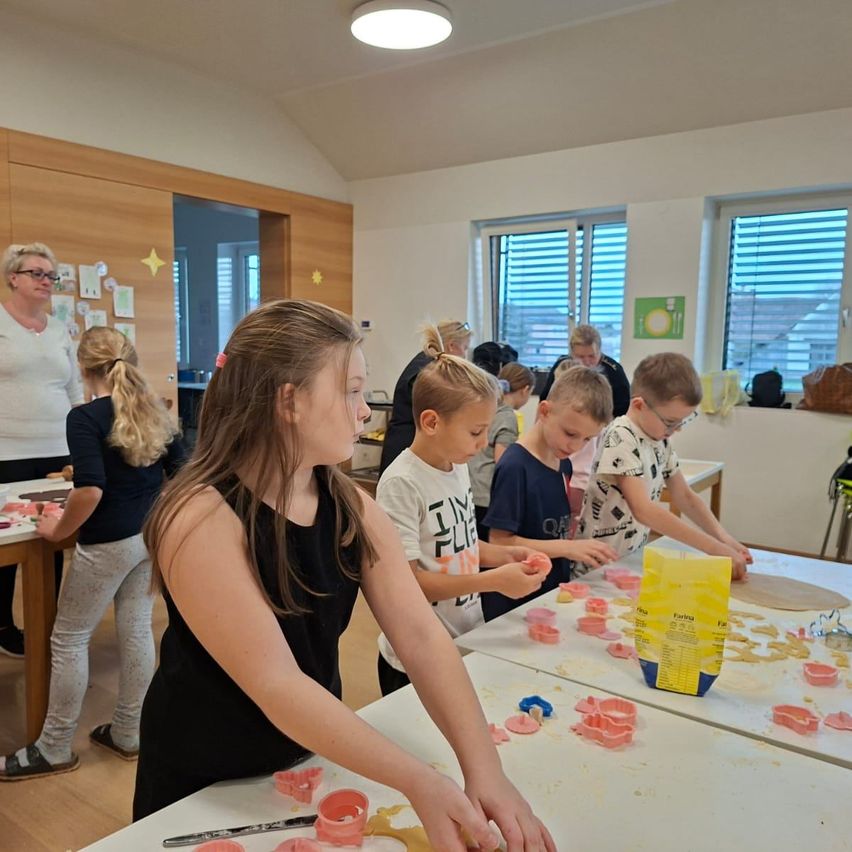 Mehrere Kinder und eine erwachsene Frau befinden sich in einem Raum und beschäftigen sich mit einer Aktivität. Sie stellen Handwerk mit Ausstechformen und Teig her. Der Raum ist gut beleuchtet, mit Holzböden und weißen Wänden. Es gibt Tische mit Teig und Ausstechformen darauf.