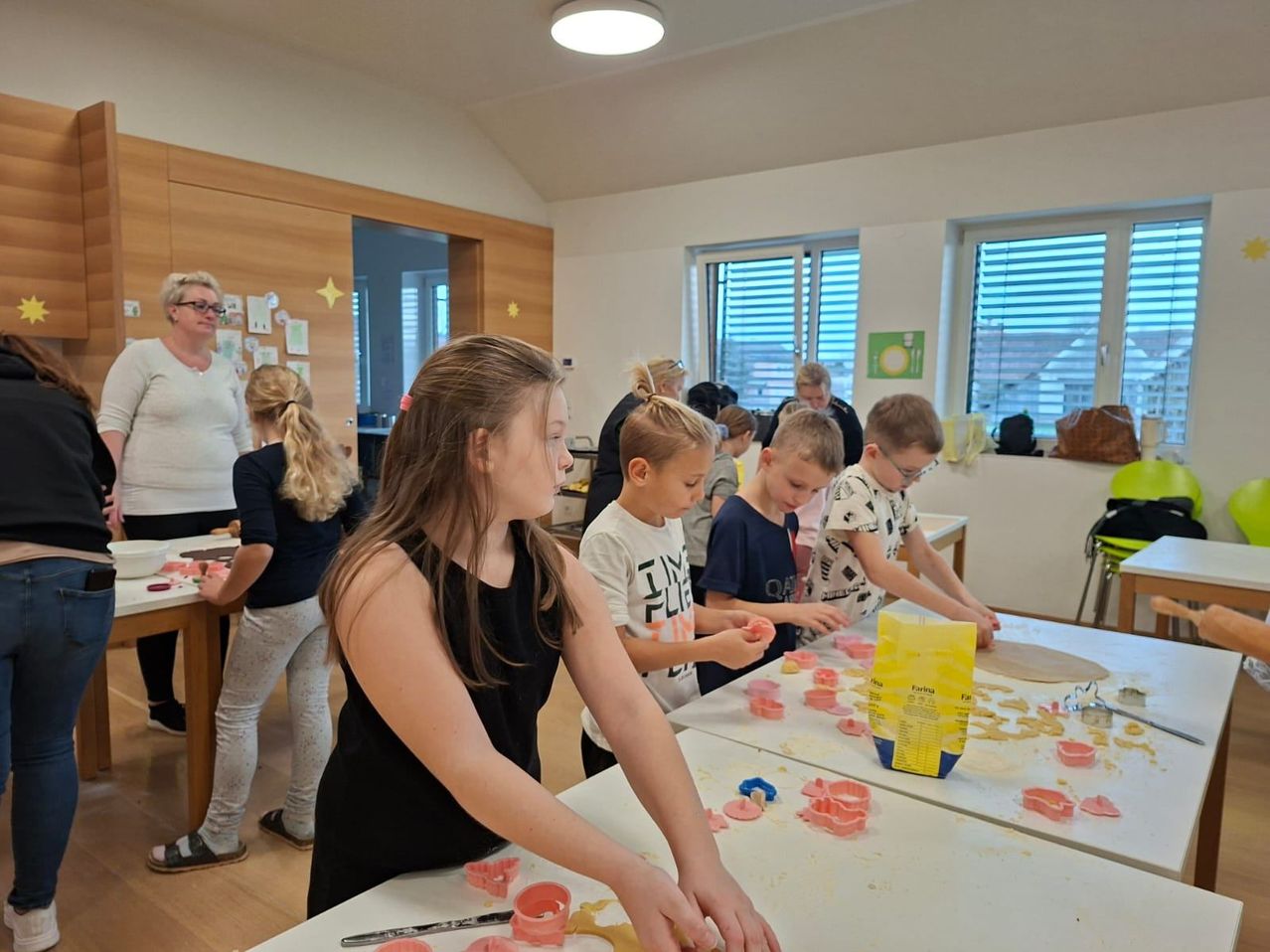 Mehrere Kinder und eine erwachsene Frau befinden sich in einem Raum und beschäftigen sich mit einer Aktivität. Sie stellen Handwerk mit Ausstechformen und Teig her. Der Raum ist gut beleuchtet, mit Holzböden und weißen Wänden. Es gibt Tische mit Teig und Ausstechformen darauf.