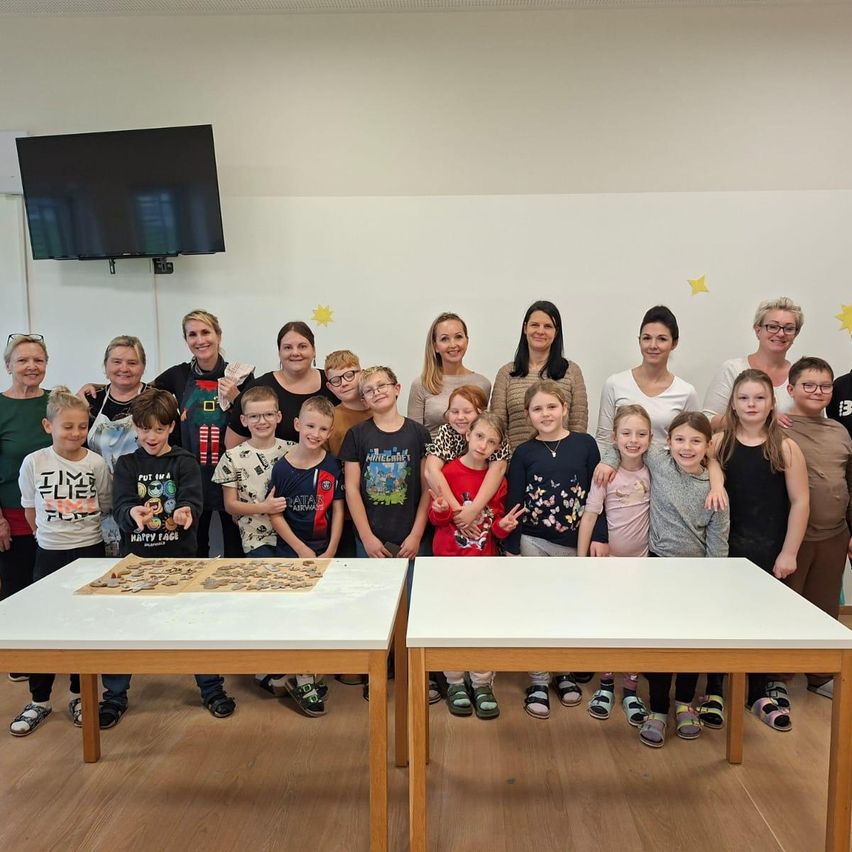 Eine Gruppe von Erwachsenen und Kindern, darunter einige mit Brille, posiert für ein Foto in einem Raum mit Holzböden und Tischen.